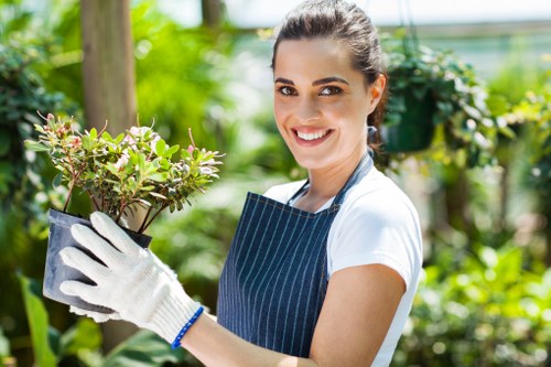 A woman smiling in a garden or greenhouse setting, wearing a navy blue apron and white gloves, holding a small potted shrub with green and pinkish leaves. The background features lush greenery, including trees and flowering plants, with a wooden post visible on the left side. The garden appears well-maintained, with a mix of natural soil, grass, and paved or decking areas, under natural daylight, suggesting a mild or sunny weather condition. The scene reflects outdoor gardening activities, highlighting plant care and landscaping work often associated with professional gardening services in the Morden area or broader London suburbs, consistent with Hedge Trimming Morden's service focus.
