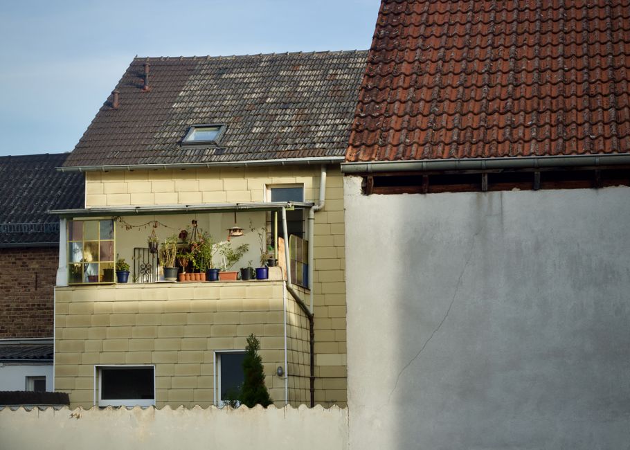 A close-up view of two residential buildings showcasing different exterior wall textures and roofing styles. The left building features a small balcony with a glass enclosure, decorated with potted pl