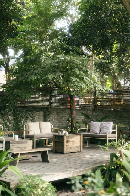A lush garden scene featuring a wooden deck with outdoor seating, including two cushioned armchairs with striped and plain cushions, and a matching wooden coffee table in the center. Surrounding the d